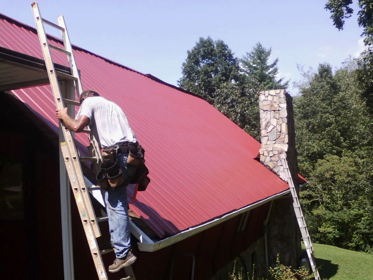 Roofer on a ladder beside a red metal roof and stone chimney — Soffit & Fascia Repair by Roofing Brothers in Sunland